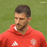 a man wearing a red manchester united hoodie is standing on a soccer field .