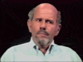 a bald man with a beard wearing a white shirt looks at the camera