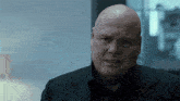 a bald man wearing a black suit looks at the camera
