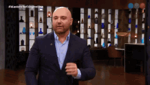 a bald man in a suit stands in front of a wall of vases with the hashtag masterchef argentina on the screen