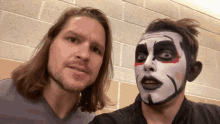 a man with long hair stands next to a man with face paint on his face