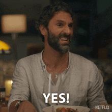 a man with a beard is sitting at a table with a bowl of food and says yes