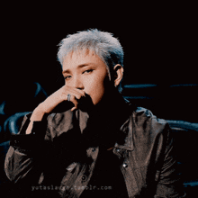 a man in a leather jacket is sitting in a dark room with his hand on his chin