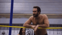 a shirtless wrestler leans on a yellow rope with the letters kj on the bottom right