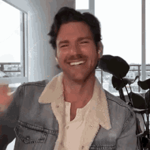 a man wearing a denim jacket and ear buds is smiling and waving at the camera .