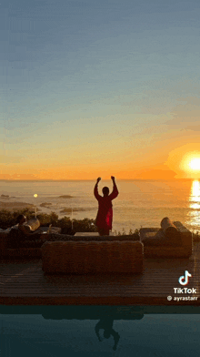 a woman in a red robe is standing on a deck overlooking the ocean at sunset .