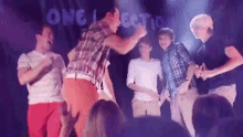 a group of men are dancing on a stage in front of a crowd with the word one direction behind them .