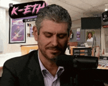 a man in a suit is sitting in front of a microphone with a sign above him that says k-eth