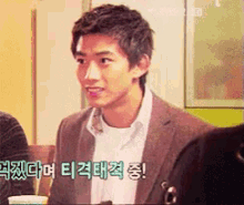 a man in a suit and white shirt is sitting at a table with korean writing on the wall behind him