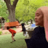 a woman with pink hair is standing in a park looking at a boy playing in the grass .
