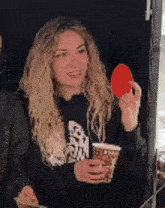 a woman in a black hoodie is holding a cup of coffee and a red heart in her hand .