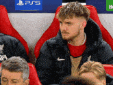 a man wearing a nike jacket sits in a stadium watching a soccer game