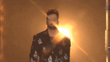 a man with a beard wearing a sequined jacket is standing in front of a wall with the sun shining through it