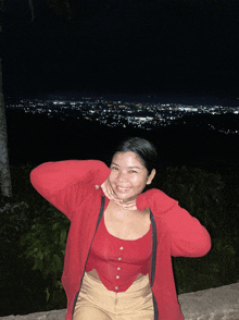 a woman wearing a red top and a red jacket stands in front of a city skyline at night