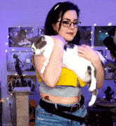 a woman wearing glasses holds a cat in her arms in front of a wall with pictures on it
