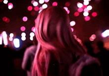 a woman with long pink hair stands in front of a blurred background of pink lights