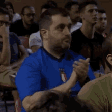 a man in a blue shirt is sitting in a crowd of people and clapping .