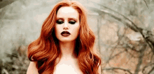 a woman with long red hair and dark makeup is standing in a forest .