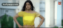 a woman in a yellow top is standing with her hands on her hips and looking at the camera .