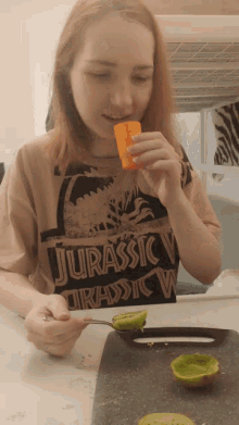 a girl wearing a shirt that says jurassic v is eating a kiwi