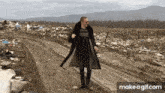 a man in a trench coat is walking down a dirt road in front of a pile of trash