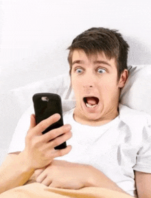a young man is laying in bed looking at his cell phone with a surprised look on his face .