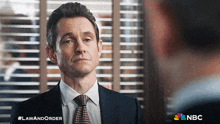 a man in a suit and tie is standing in front of a window with the nbc logo in the corner