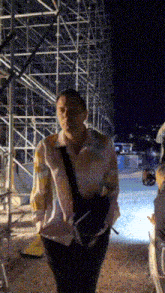 a man in a striped shirt is walking in front of a scaffolding at night .