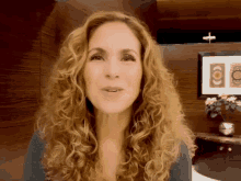 a woman with curly hair looks at the camera in front of a picture of a cross