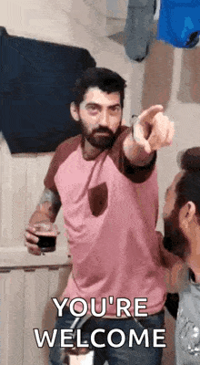 a man with a beard is pointing at another man while holding a glass of beer and saying you 're welcome .