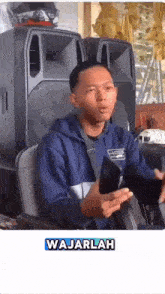 a man in a blue jacket is holding a cell phone in front of speakers and the word wajarlah is on the bottom