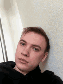 a young man leans against a white wall and looks at the camera with a serious look on his face