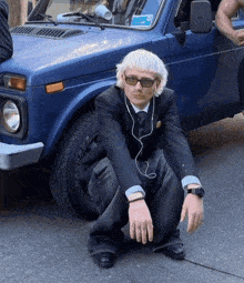 a man squatting in front of a blue car with a parking ticket