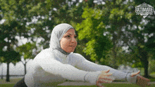 a woman wearing a hijab is stretching her arms in a park