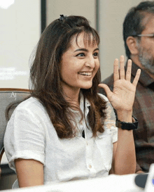 a woman in a white shirt is smiling and waving at the camera