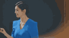 a woman in a blue dress is standing in front of a black wall and holding a cell phone .
