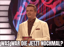 a man in a white suit and bow tie is holding a piece of paper with the words was war die jetzt nochmal on it