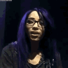 a woman with purple hair and glasses is wearing a black jacket .