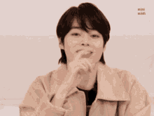 a young man in a tan jacket is smiling with his hands on his chin .
