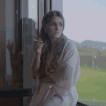 a woman in a robe is smoking a cigarette while looking out a window
