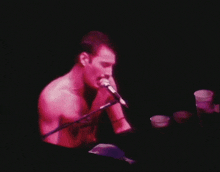 a shirtless man singing into a microphone with a pink light behind him