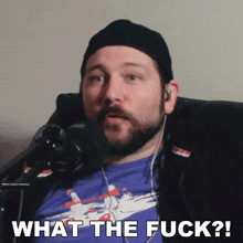 a man with a beard is sitting in front of a microphone with the words what the fuck written below him