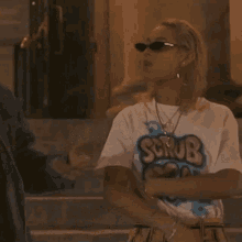 a woman wearing sunglasses and a white shirt that says scrub on it