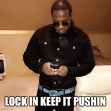 a man wearing sunglasses looks at his phone with the caption lock in keep it pushin below him