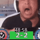 a man wearing headphones is yelling into a microphone while a scoreboard displays the score 2-2