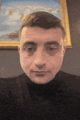a man in a black turtleneck looks at the camera in front of a framed painting