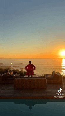 a woman in a red robe is standing in front of a sunset over the ocean .