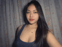 a woman wearing a blue tank top and a necklace stands in front of a patterned curtain