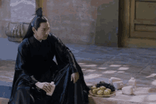a man in a black robe sits next to a plate of grapes