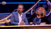 a man and a woman are sitting at a table in front of a masked singer logo